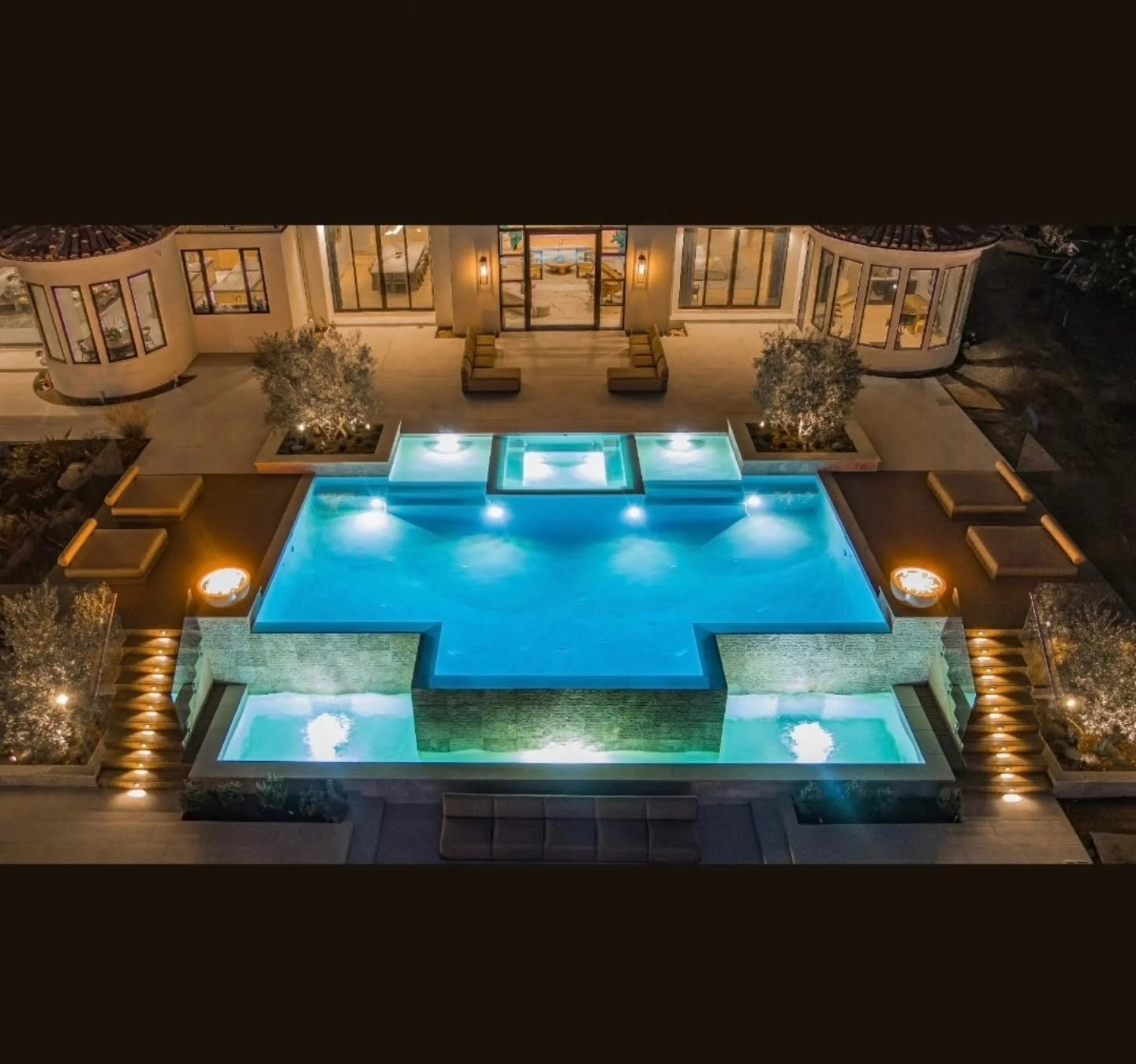 Aerial view of illuminated pool deck with stone hardscape, spa features, and integrated landscape lighting