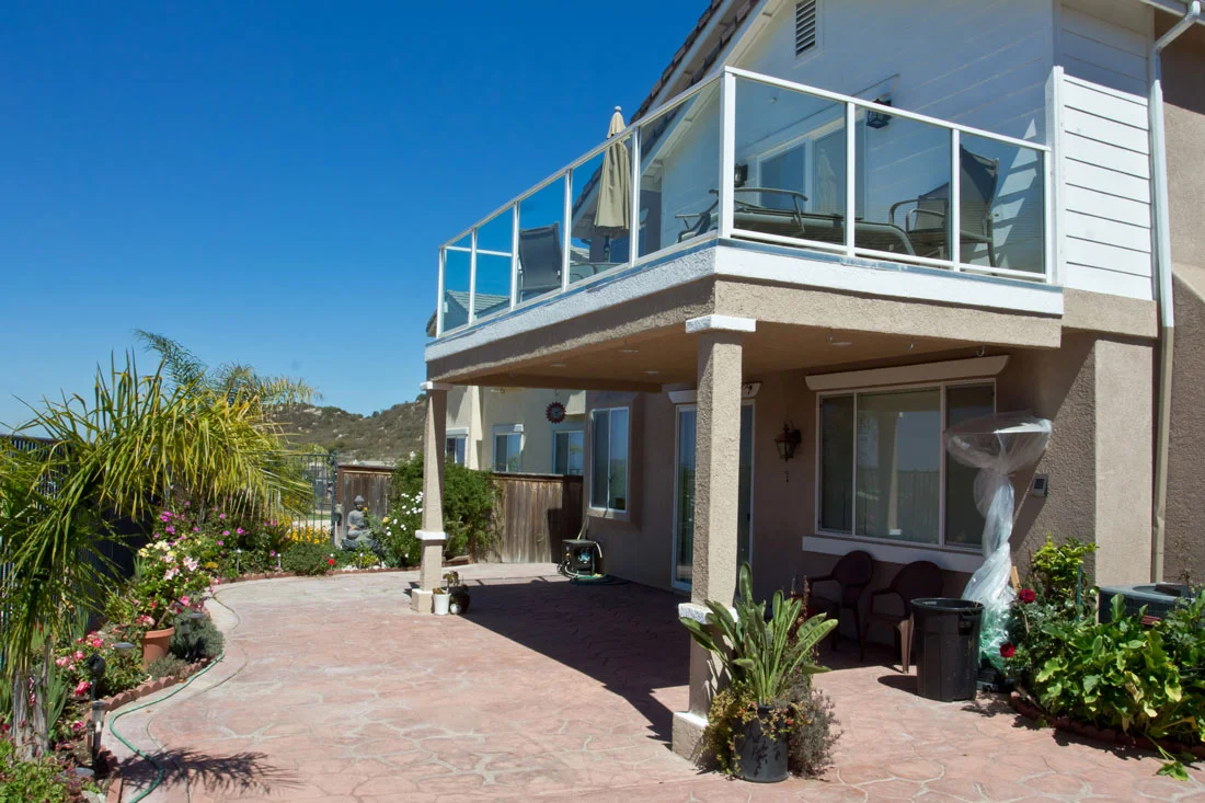 Elevated white deck with glass railings overlooking terracotta patio and covered structure below